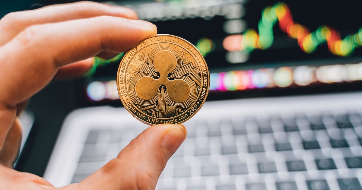 A close-up view of a hand holding a Ripple cryptocurrency coin with a blurred computer in the background.
