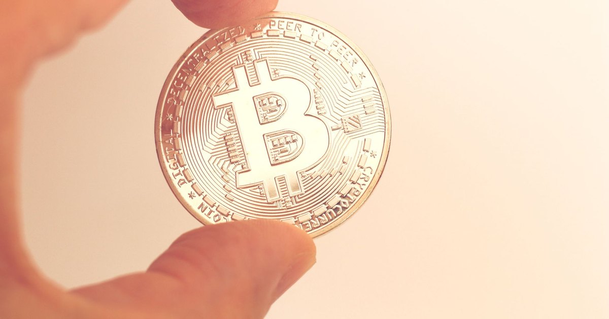 A close-up image of a hand holding a Bitcoin coin against a neutral background.