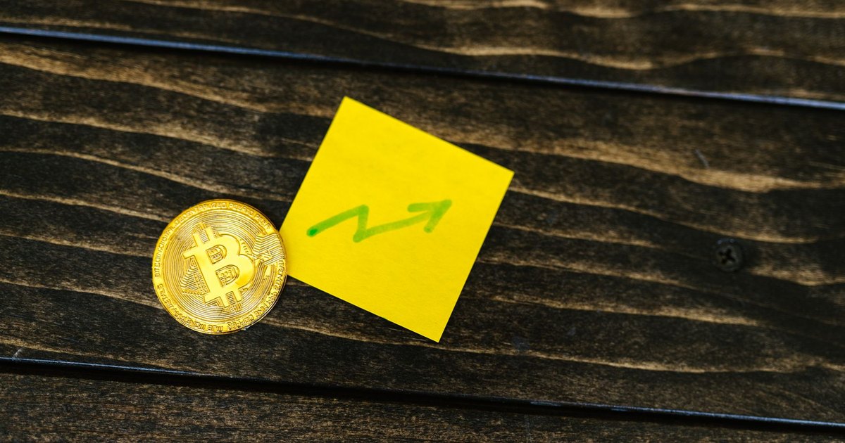 Close-up of a Bitcoin coin with an upward arrow sticky note on a wooden surface.