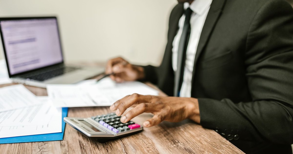 A businessman in a black suit using a calculator at a desk with financial documents and a laptop.