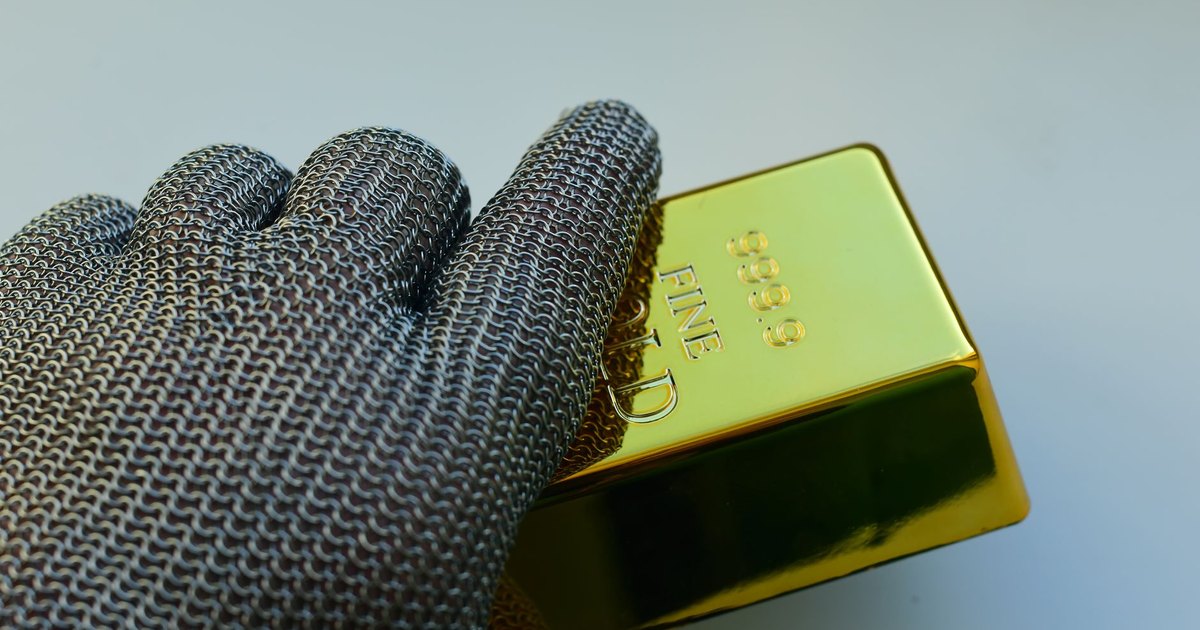 Close-up of a metal gloved hand holding a shiny gold bar indoors.