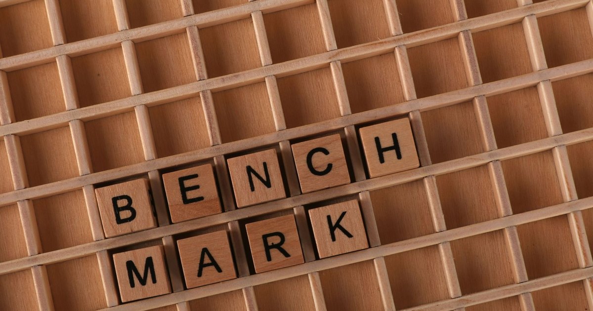 Top view of wooden letter tiles arranged to spell 'Benchmark' on a wooden grid.