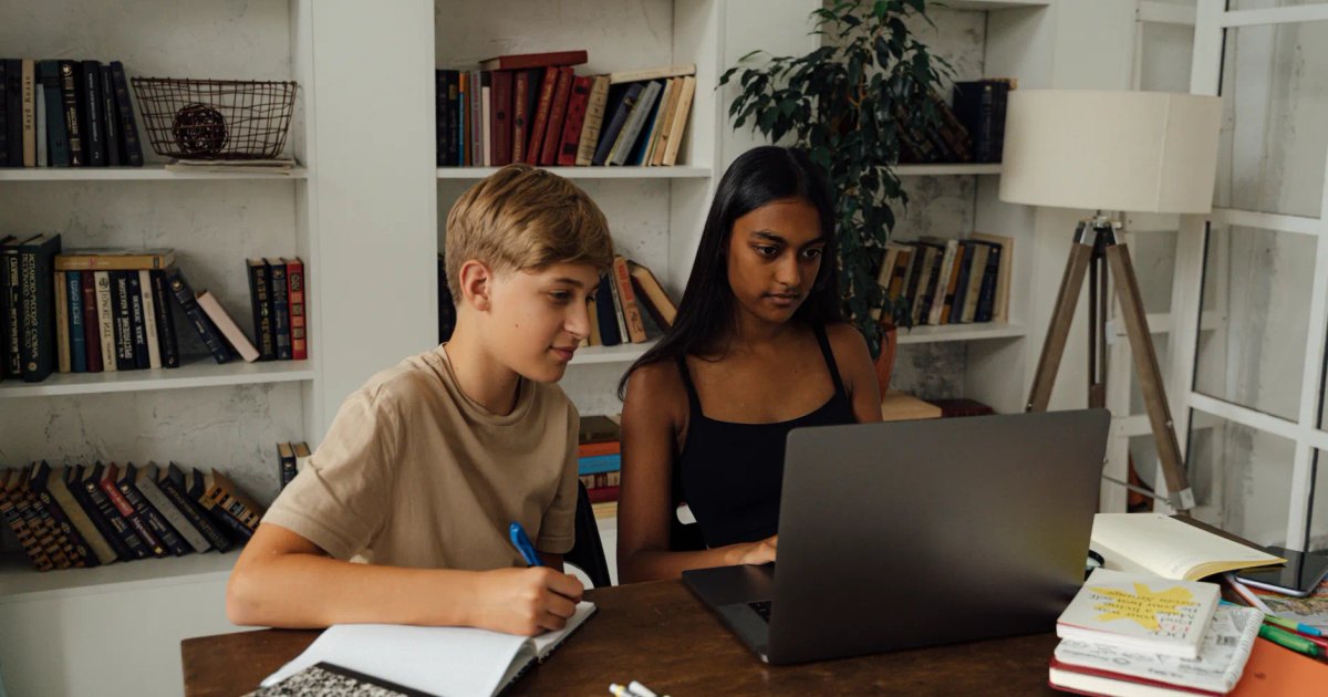 A boy and girl study together using a laptop in a home library setting.